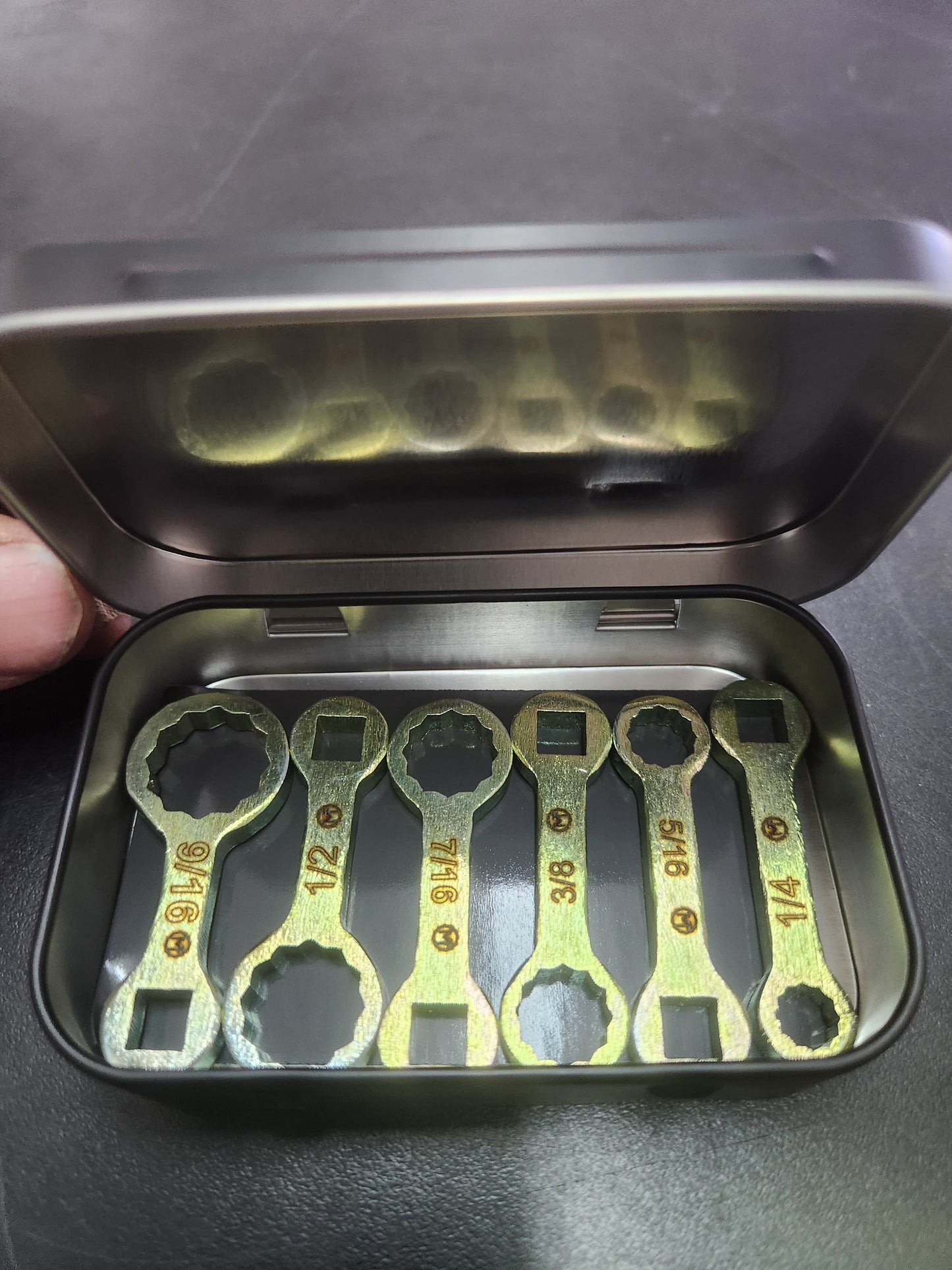 A set of six gold-colored dog bone-shaped wrenches in a variety of sizes, displayed in an open metal tin.