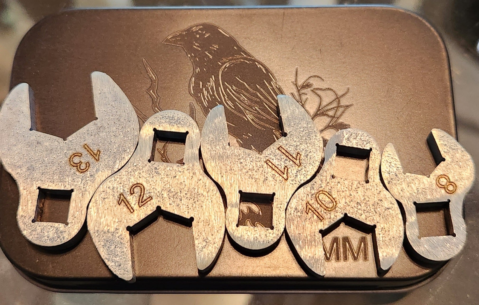 A set of five metal crows foot mm wrenches in increasing sizes, placed side by side on a metal surface with a crow silhouette behind them.